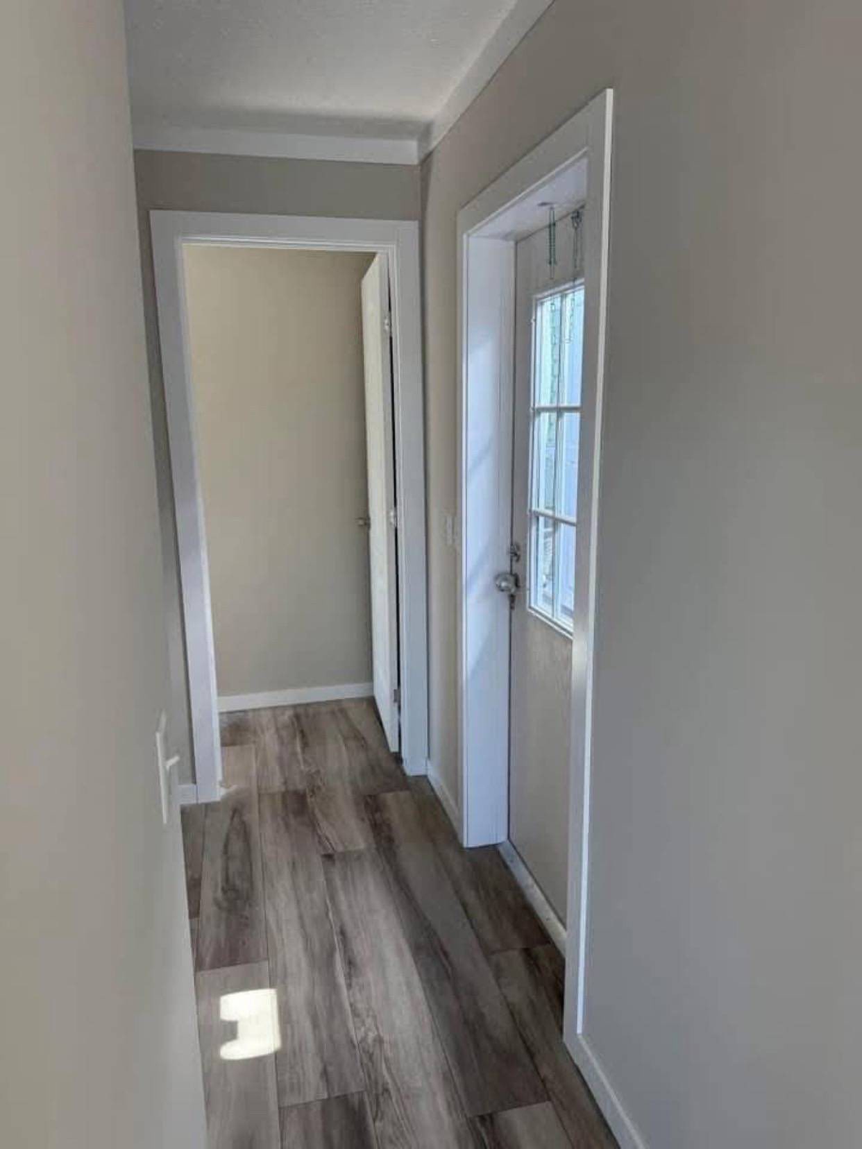 Narrow hallway with light gray walls, wood flooring, a white-framed door on the left, and a glass door on the right. Bright natural light streams in, creating a calm ambiance.