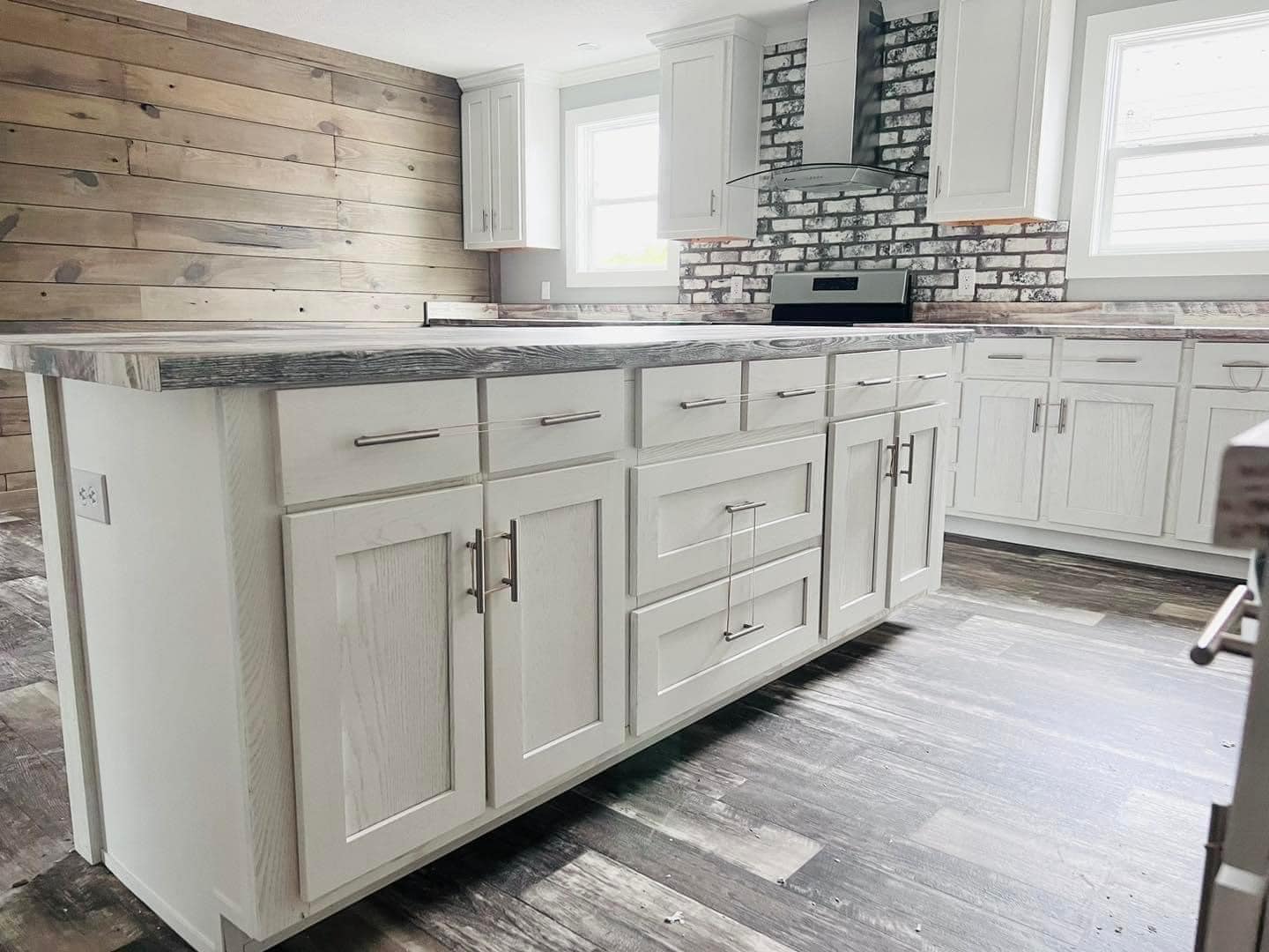 Modern kitchen with a rustic flair, featuring white cabinets, a gray wooden island, brick backsplash, and wood-paneled walls. Bright and airy atmosphere.