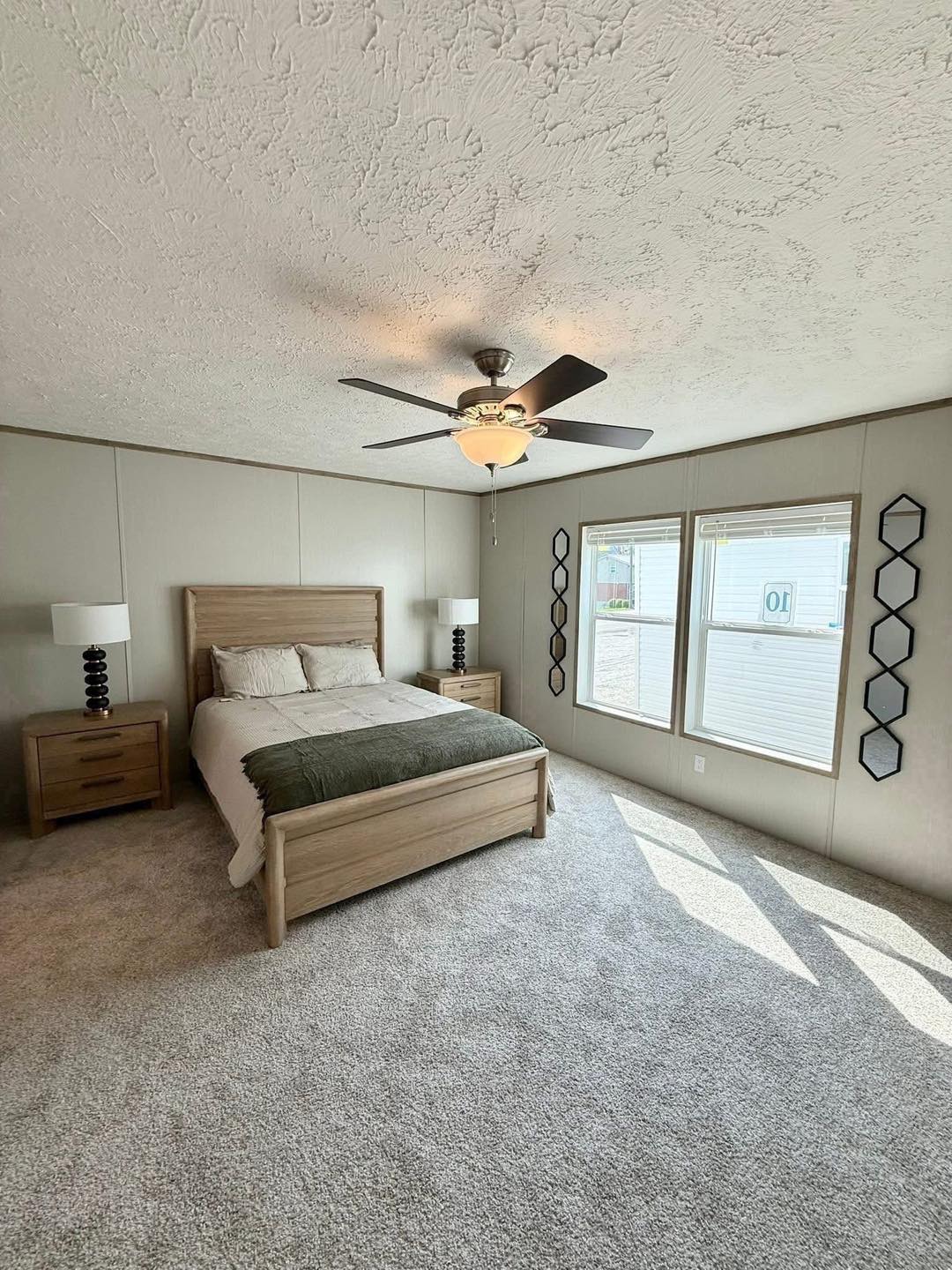 A modern bedroom with a wooden bed, white and green bedding, flanked by two nightstands with lamps. A ceiling fan above and natural light from two windows.