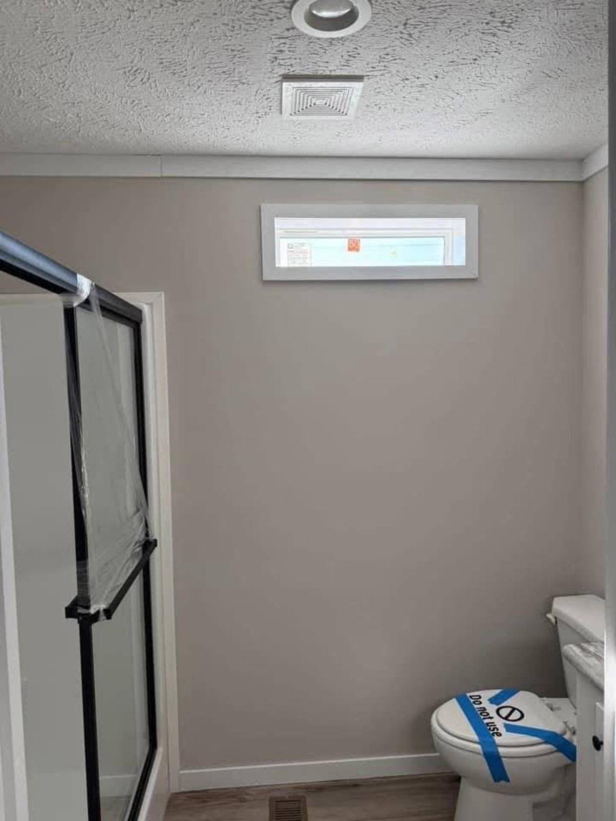 Small bathroom with gray walls, ceiling light, and a window. Shower with a glass door on the left, toilet with “Do Not Use” tape on the right.