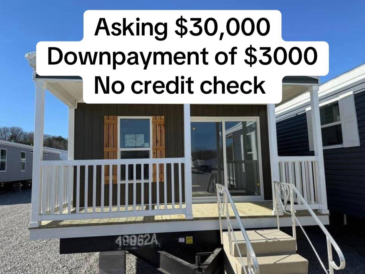 Small, dark-paneled mobile home with white railing, set in a lot. Text overlay reads "Asking $30,000, Downpayment of $3,000, No credit check." Bright, sunny day.