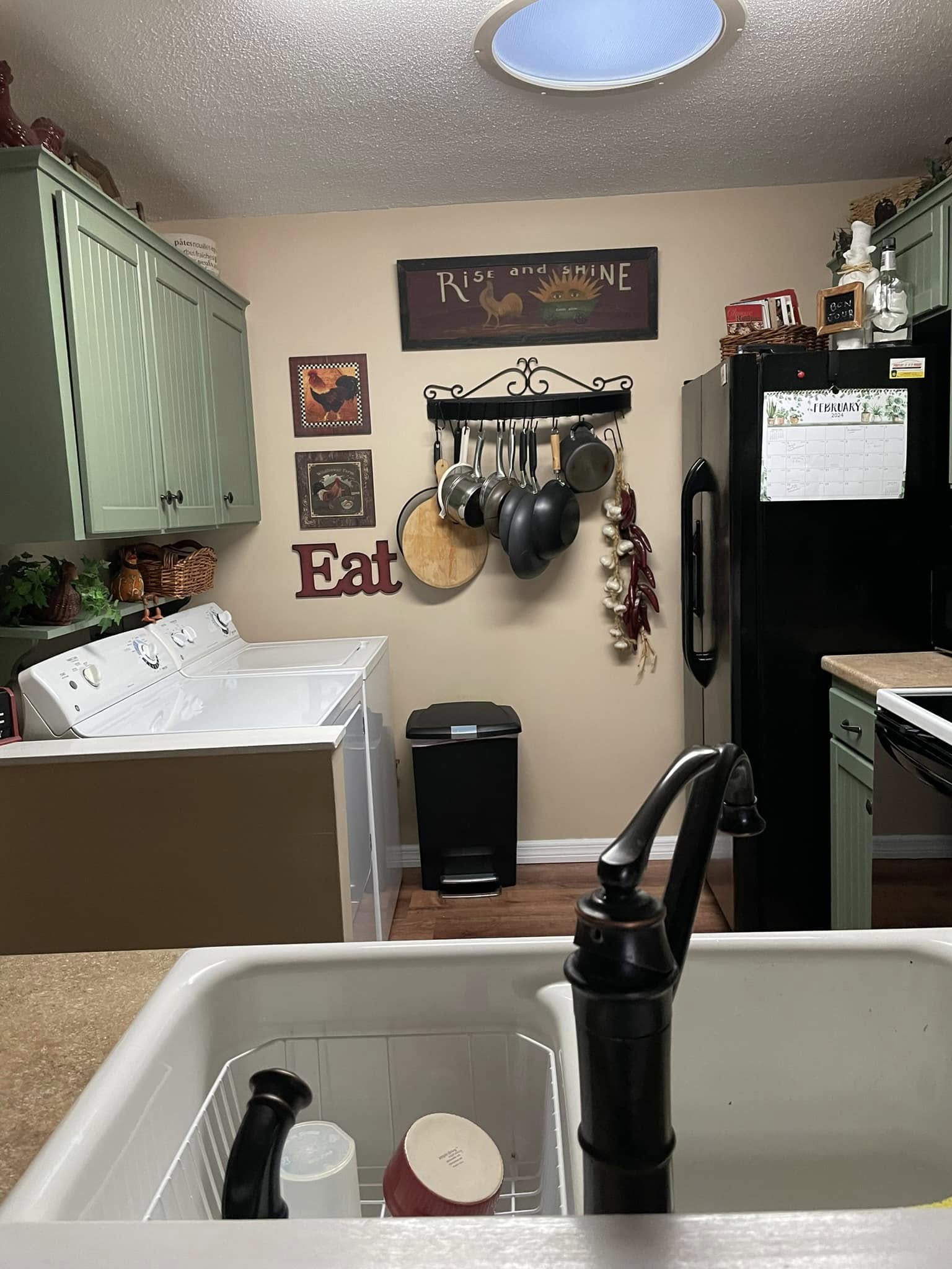 Cozy kitchen with green cabinets, black fridge, and wall art. Pots hang on rack, "Eat" sign decorates wall. Washer/dryer in corner, creating a homely vibe.