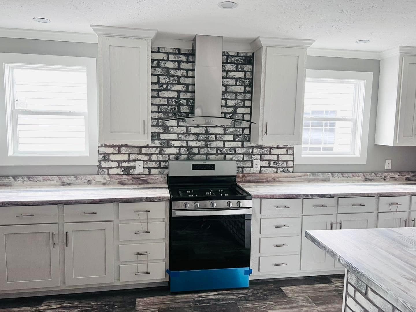 Modern kitchen with a stainless steel stove centered between two white cabinets against a white brick backsplash. Light streams through two windows.
