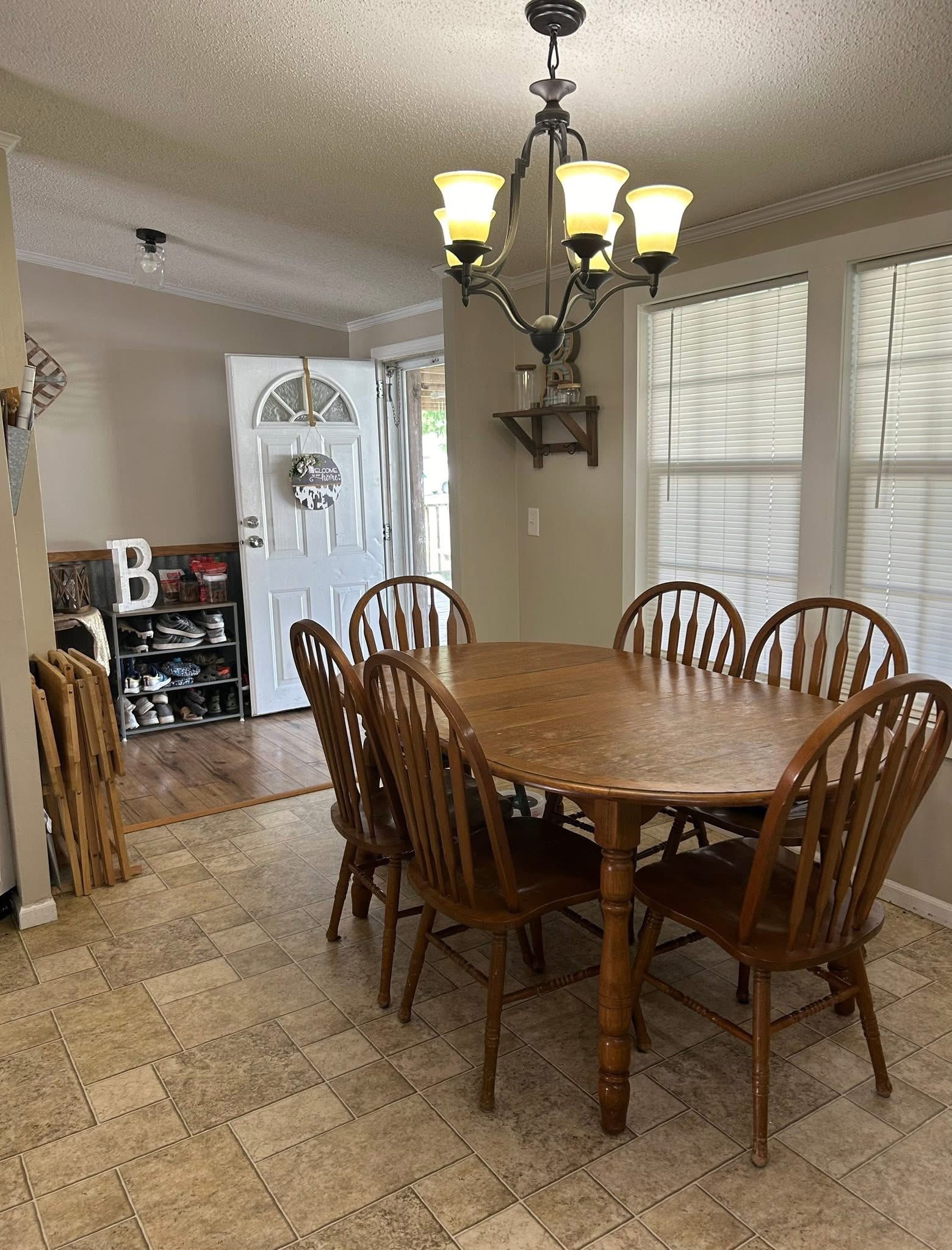 Dining room with a wooden table and six chairs, under an elegant chandelier. Door is open, showing a shoe rack and a welcoming ambiance.