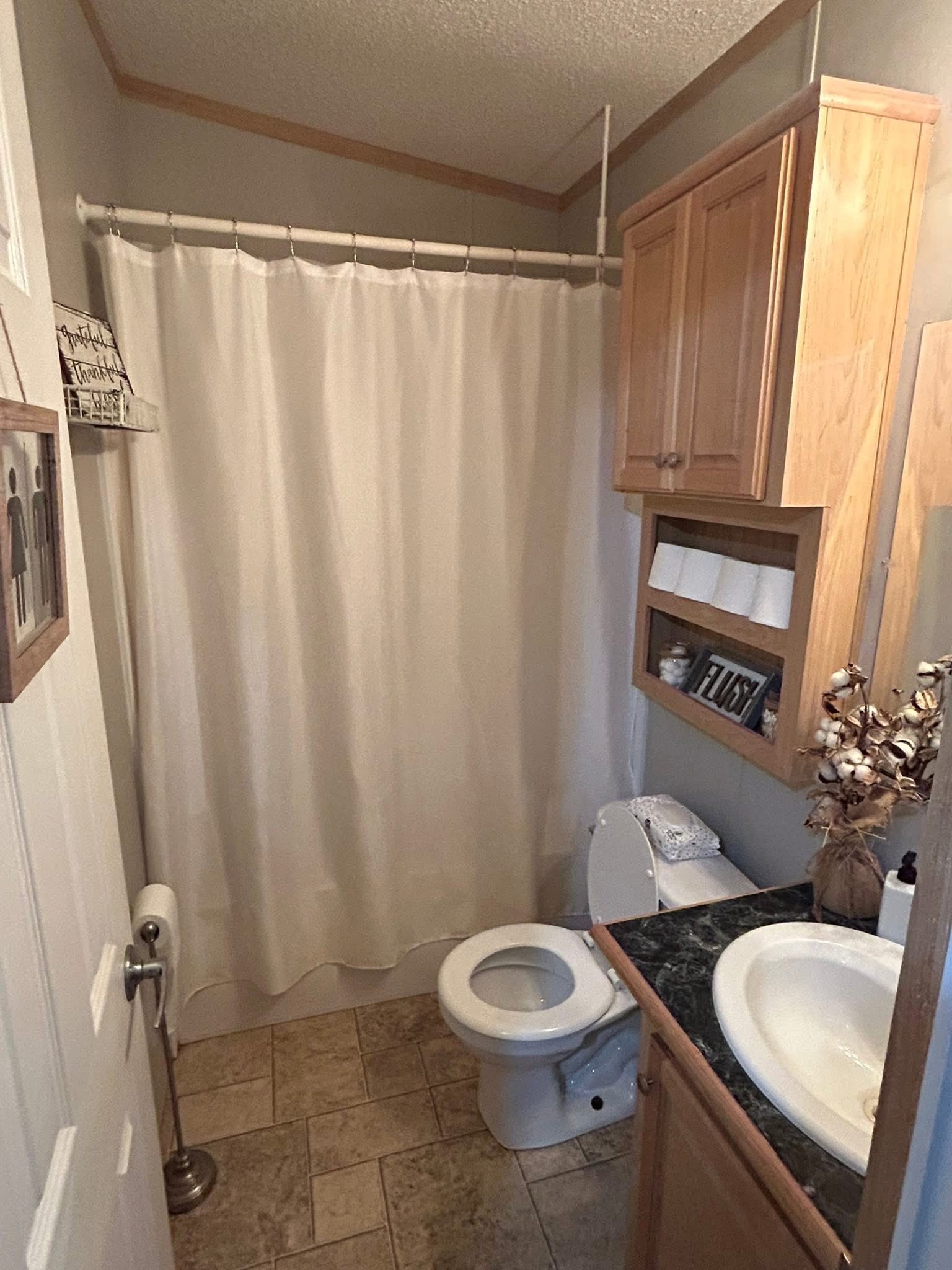 Compact bathroom with beige tiles and gray walls. Features a white shower curtain, toilet, sink with a marble countertop, and wooden storage cabinets.