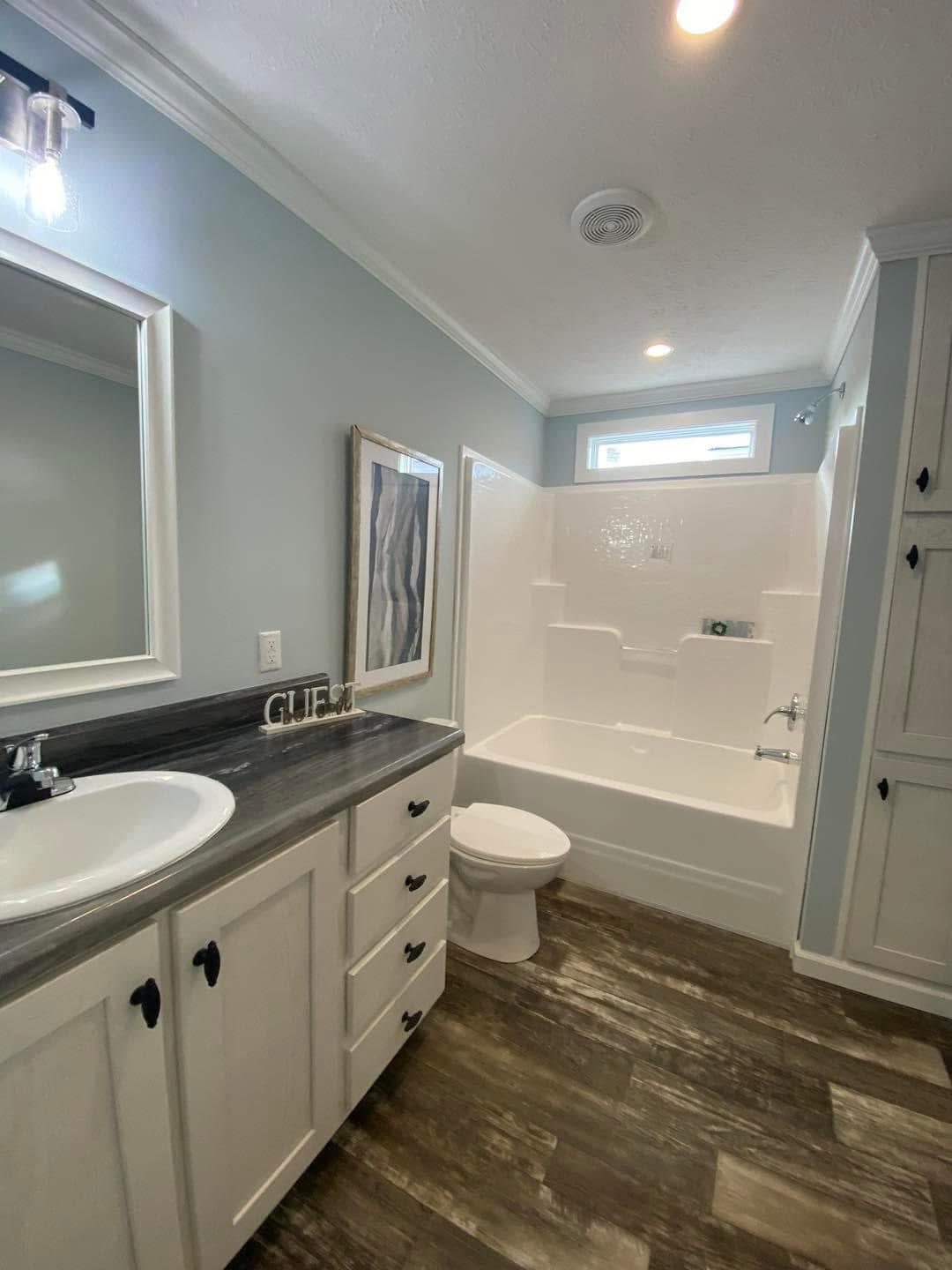 A modern bathroom with light blue walls, featuring a white vanity, dark countertop, and circular sink. The bathtub is white, next to a toilet. The floor has a wood finish, creating a clean and inviting atmosphere.