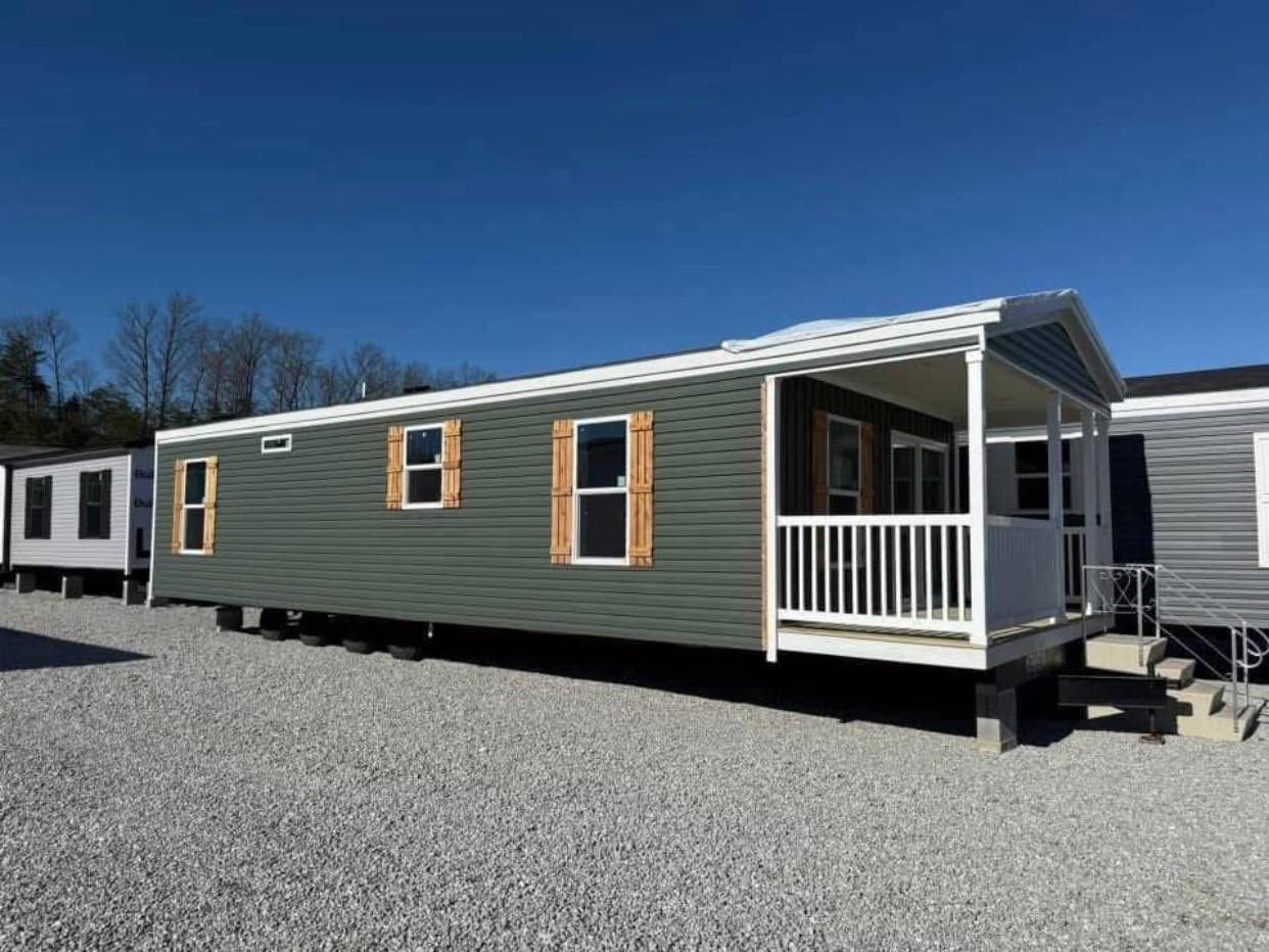A green mobile home with wooden shutters and a white-railed porch sits on gravel under a clear blue sky. The setting is calm and sunny.