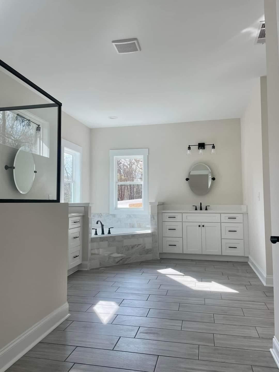 Modern bathroom with gray tile flooring, double vanity, round mirrors, and a large bathtub beside two windows. Sunlight softly illuminates the space.
