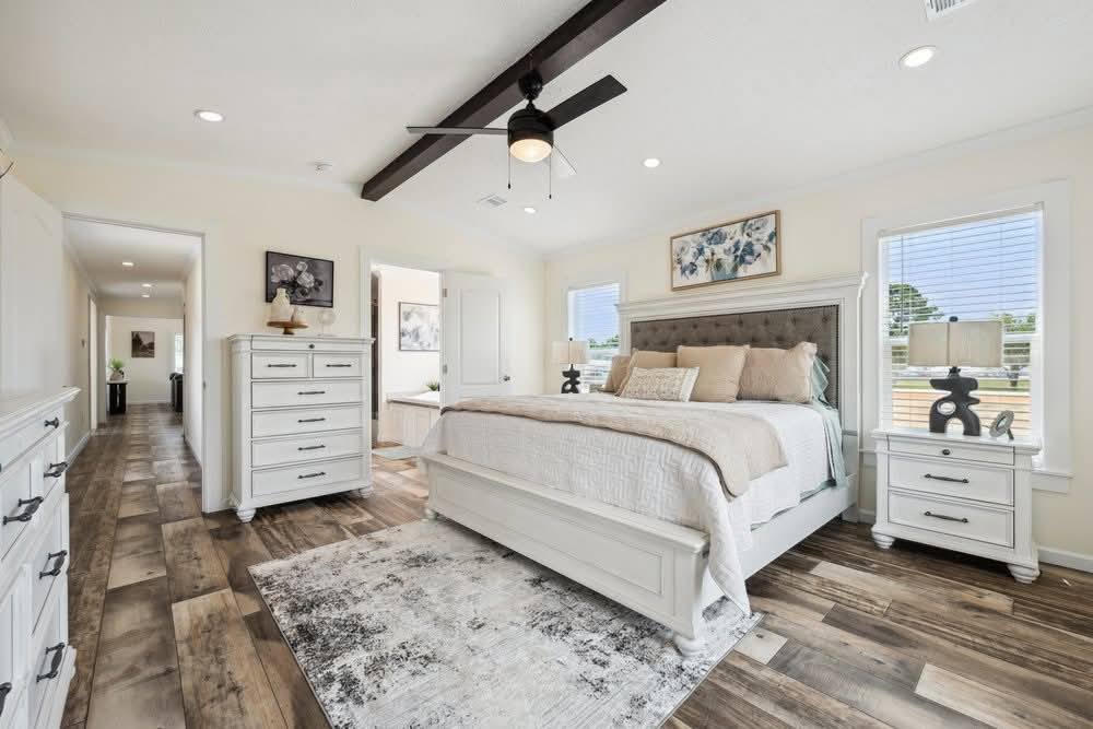Bright, elegant bedroom with a white king-size bed, wooden floor, cream accents, and ceiling fan. Two windows illuminate the peaceful setting.