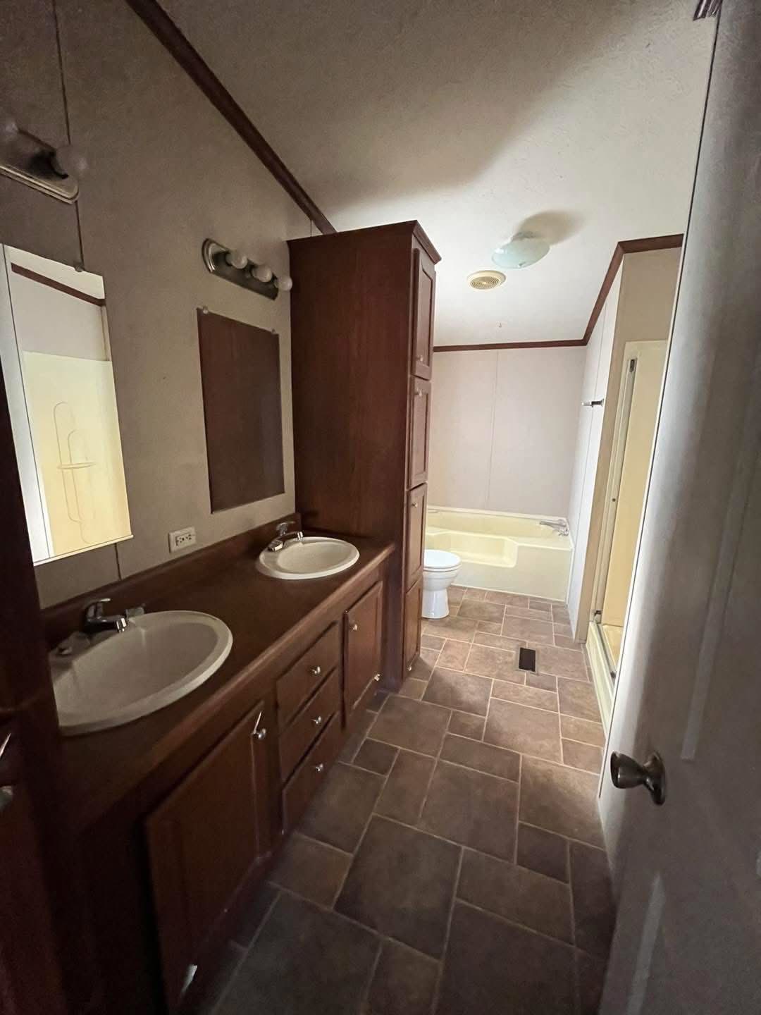 A narrow bathroom with two sinks on a wooden vanity, mirrors, and overhead lights. A bathtub, toilet, and shower are visible in the background.