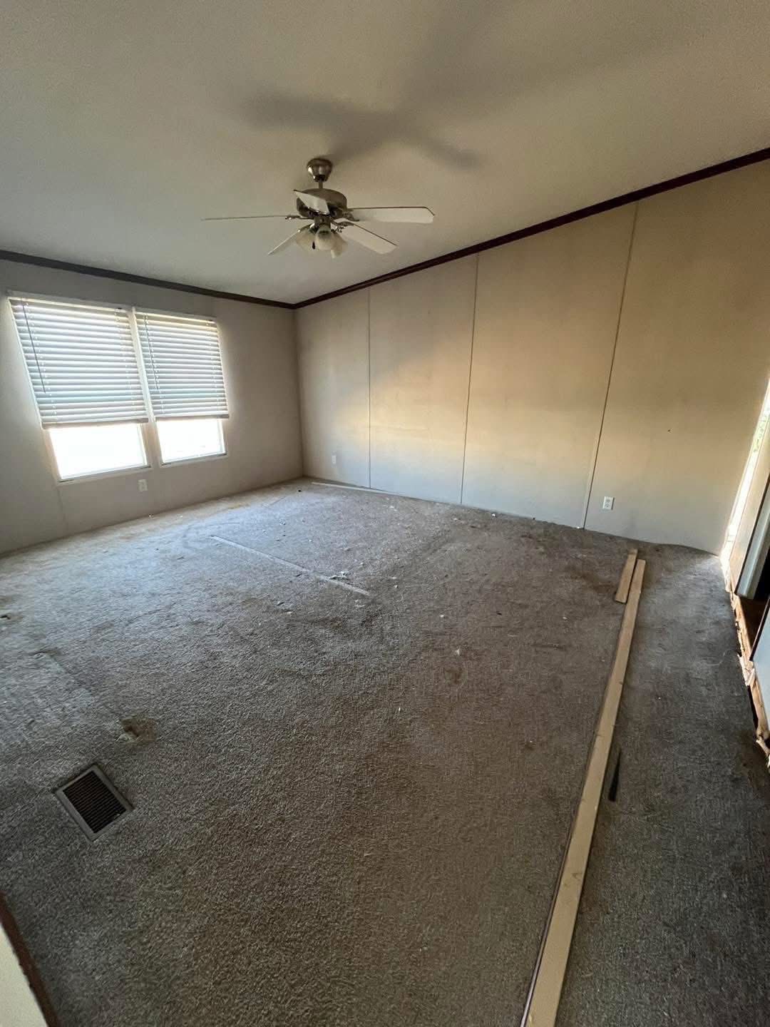 An empty, dimly lit room with beige walls and slightly stained carpet. Two windows with blinds are on one wall, and there's a ceiling fan above.