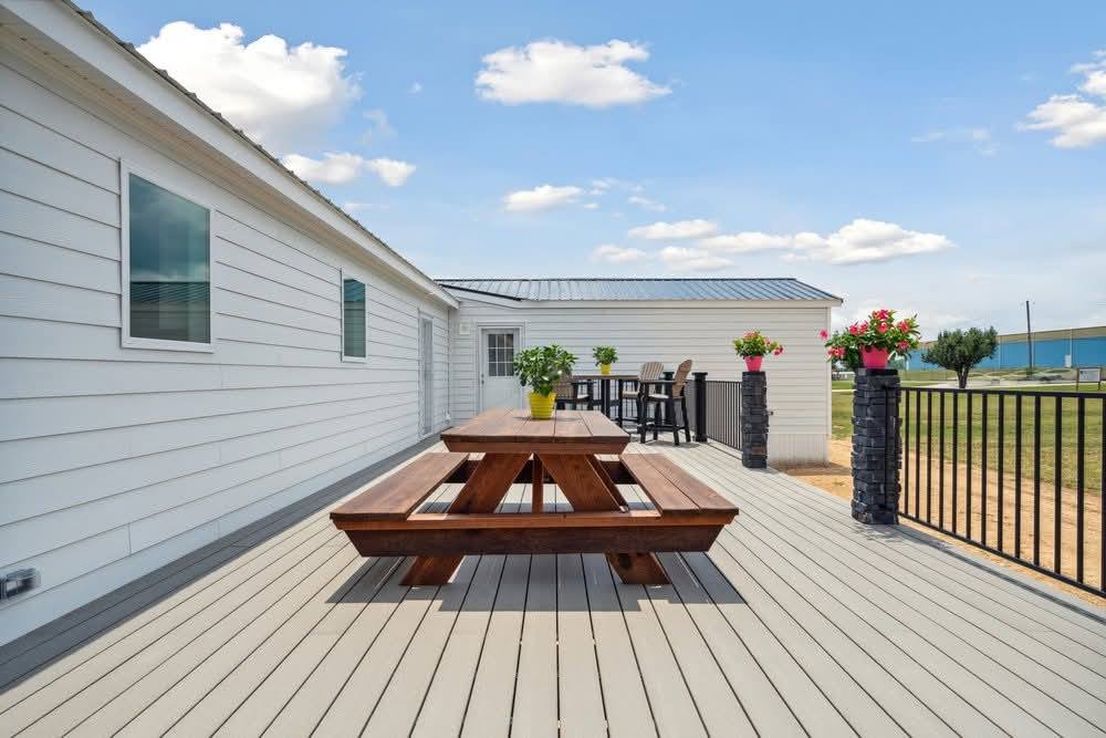 A spacious wooden deck features a picnic table with a plant in the center. Potted flowers line one side. The scene is bright and open under a clear blue sky.