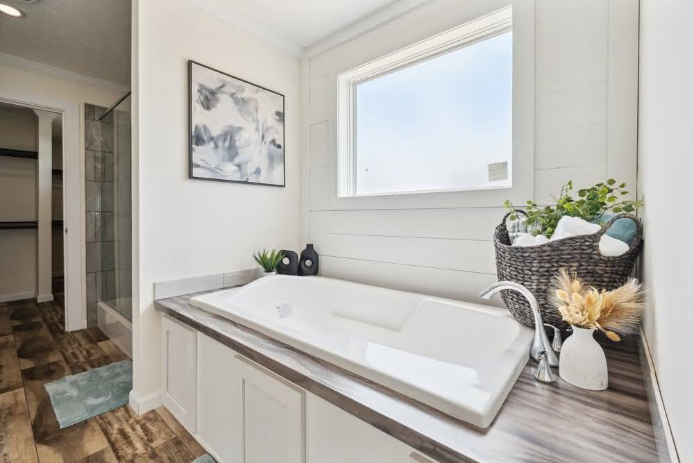 A modern bathroom with a white, built-in bathtub under a large window. The wooden floor, abstract painting, and potted plants create a serene atmosphere.