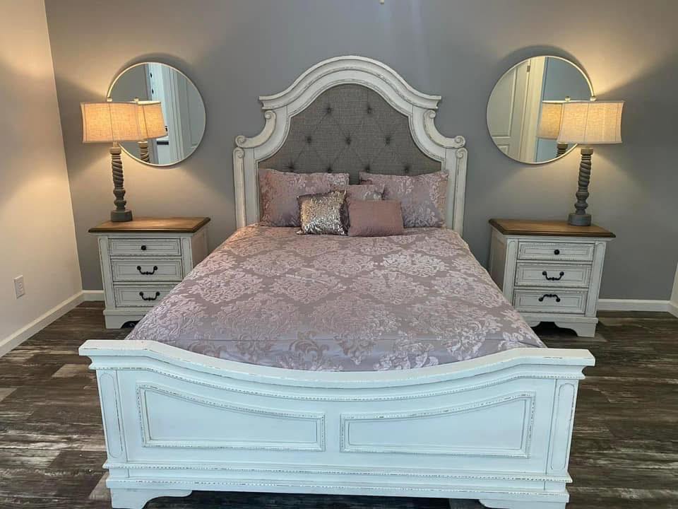 Classic bedroom with a white ornate bed, lavender bedding, and a gray tufted headboard. Flanked by round mirrors and matching lamps on nightstands.