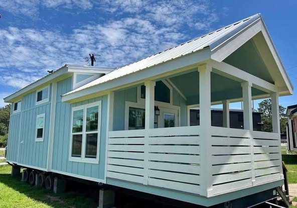 Modern tiny house on wheels, painted light blue with white trim. Features large windows and a covered porch. A sunny day with blue sky and clouds.