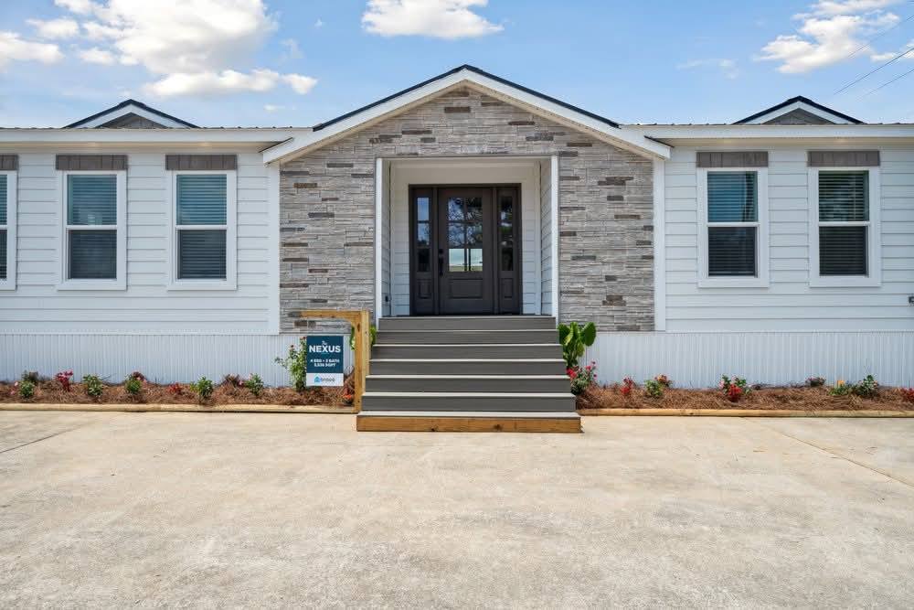 Modern single-story house with a stone and white exterior, symmetrical windows, and gray double doors. Front yard has neat landscaping and a driveway.