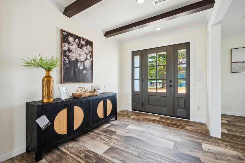 A stylish entryway with wooden flooring, a dark console with circular motifs, and a decorative vase. Double doors allow natural light, creating a welcoming ambiance.