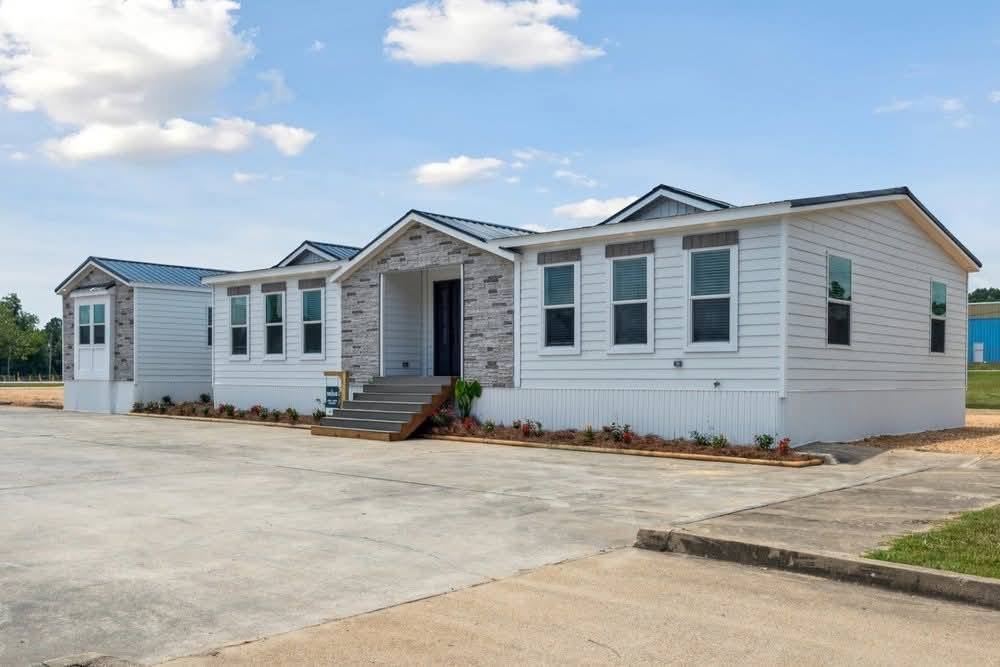 A modern white manufactured home with a stone entrance facade, multiple windows, and a neatly landscaped driveway under a clear blue sky.