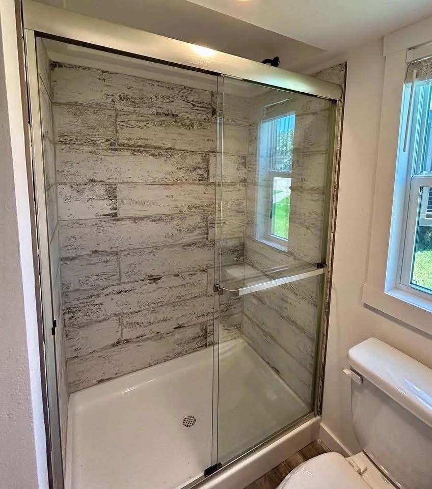 Modern bathroom interior with a rustic, wood-patterned tile shower and sliding glass doors. Natural light from a nearby window creates a serene ambiance.