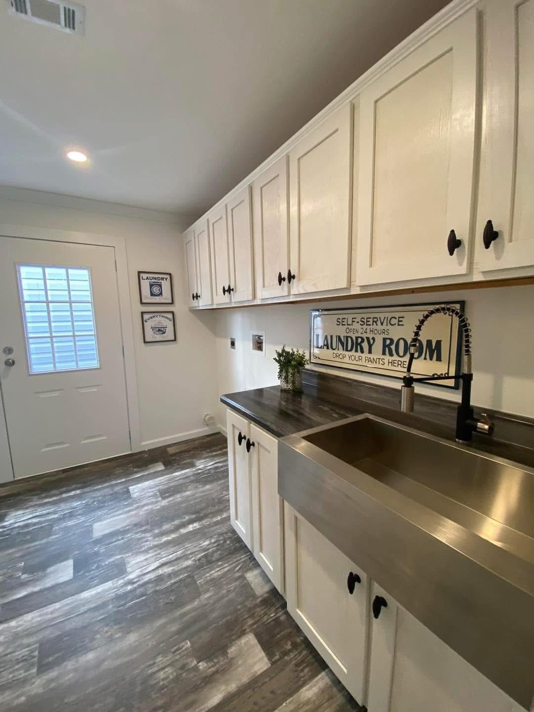Bright laundry room with white cabinets, stainless steel farmhouse sink, grey wood floor, and humorous wall sign. Clean, modern, and inviting atmosphere.