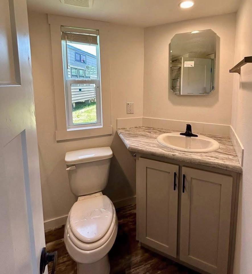 Small bathroom with a toilet and a corner sink under a modern mirror. A window reveals an outdoor view. The room has a clean, minimalist feel.