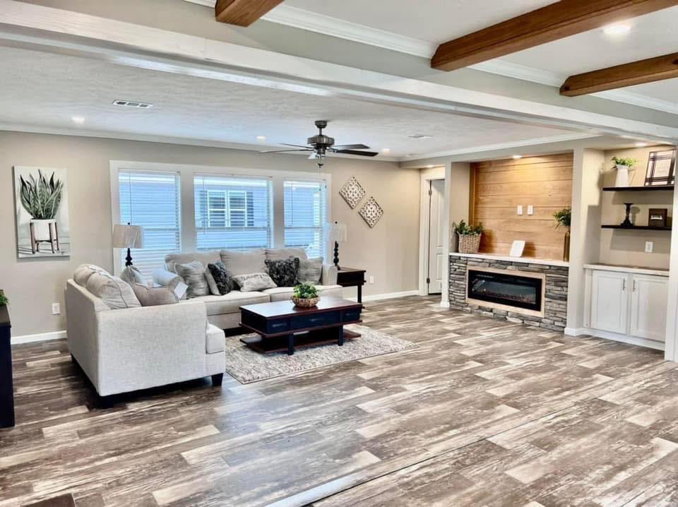 Modern living room with a beige sectional couch, wooden coffee table, large windows, ceiling fan, and a cozy electric fireplace set into a stone wall.