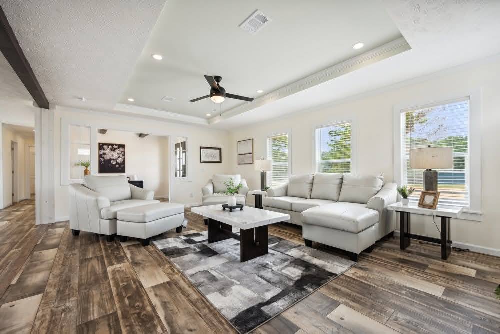 Spacious living room with white sofas and a matching armchair on a gray-patterned rug. Wooden floor, ceiling fan, lamps, and large windows adding natural light.