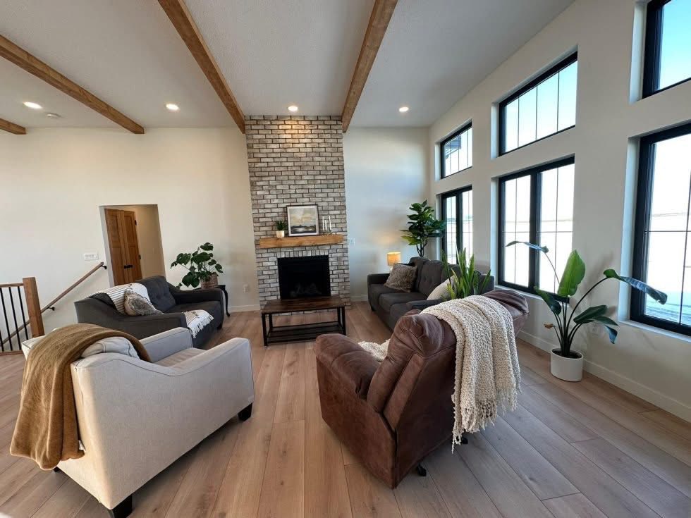 Spacious living room with large windows, brick fireplace, wooden beams, and light wooden floor. Cozy seating includes sofas and potted plants. Warm ambiance.