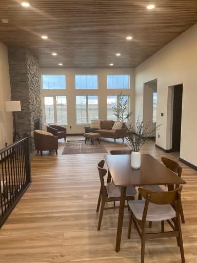 A spacious living room with a wooden ceiling and floor, stone fireplace, large windows, brown furniture, and a dining table with chairs. The room feels cozy and modern.