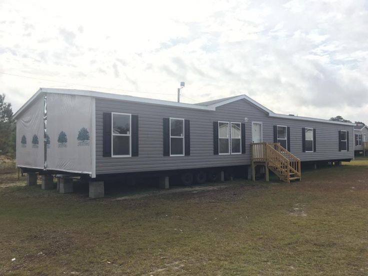 A large gray mobile home with white trim and black shutters sits on a grassy plot. A small wooden staircase leads to the front door under an overcast sky.