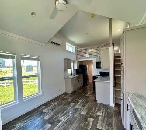 Modern tiny home interior with high ceilings, natural light from large windows, a loft area, sleek kitchen with light cabinets, and dark appliances. Cozy and airy atmosphere.