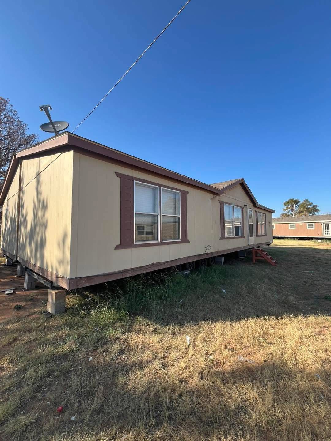 A beige mobile home with brown trim sits on a grassy lot under a clear blue sky. A satellite dish is mounted on the roof. Nearby, another mobile home is visible.
