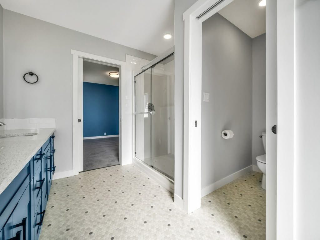 Modern bathroom with blue cabinets, white countertops, and a glass shower. Gray walls add a sleek feel, leading to a room with a blue accent wall.