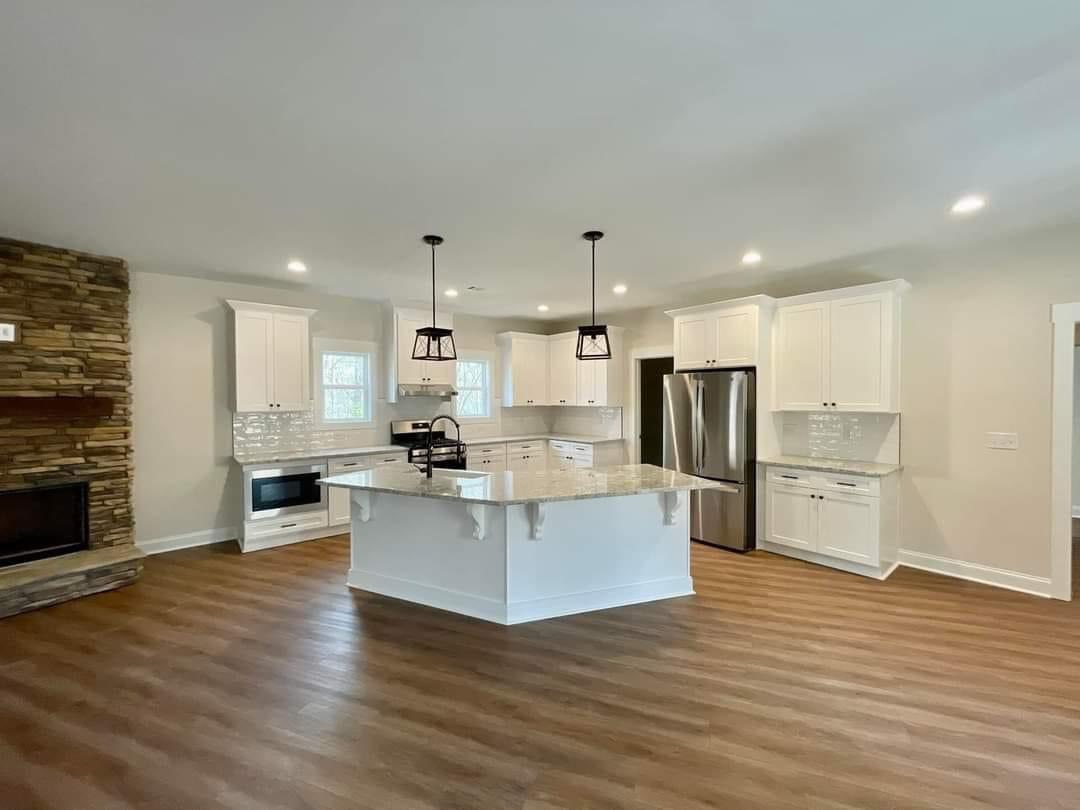 Spacious kitchen with white cabinets, a large center island with granite countertops, stainless steel appliances, wood flooring, and stone-accented fireplace.