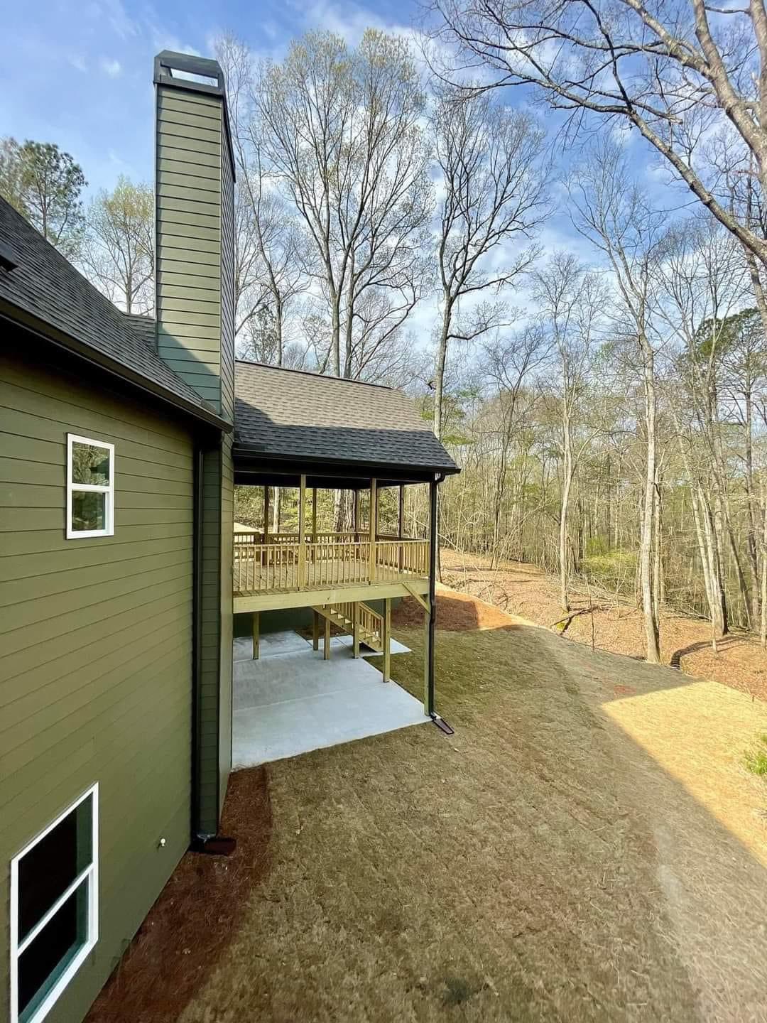 The image shows a green house with a tall chimney and a large wooden deck. Surrounding the house are leafless trees under a clear blue sky.