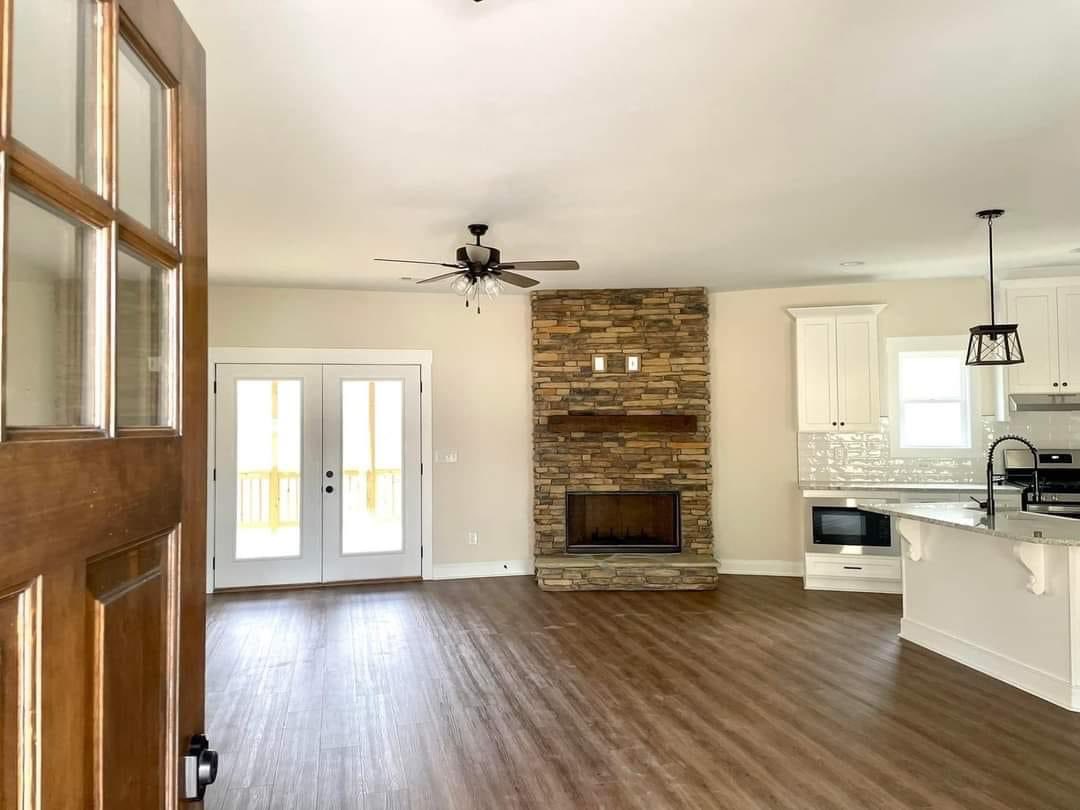 Open door view of a modern living room with a hardwood floor, stone fireplace, ceiling fan, and a bright kitchen. French doors lead outside. Cozy ambiance.