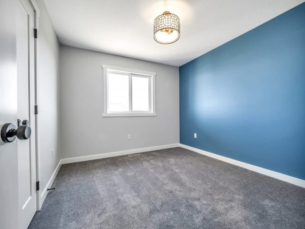 Empty room with gray carpet, white walls, and a blue accent wall. A window centered on the back wall lets in natural light. A modern ceiling light adds warmth.