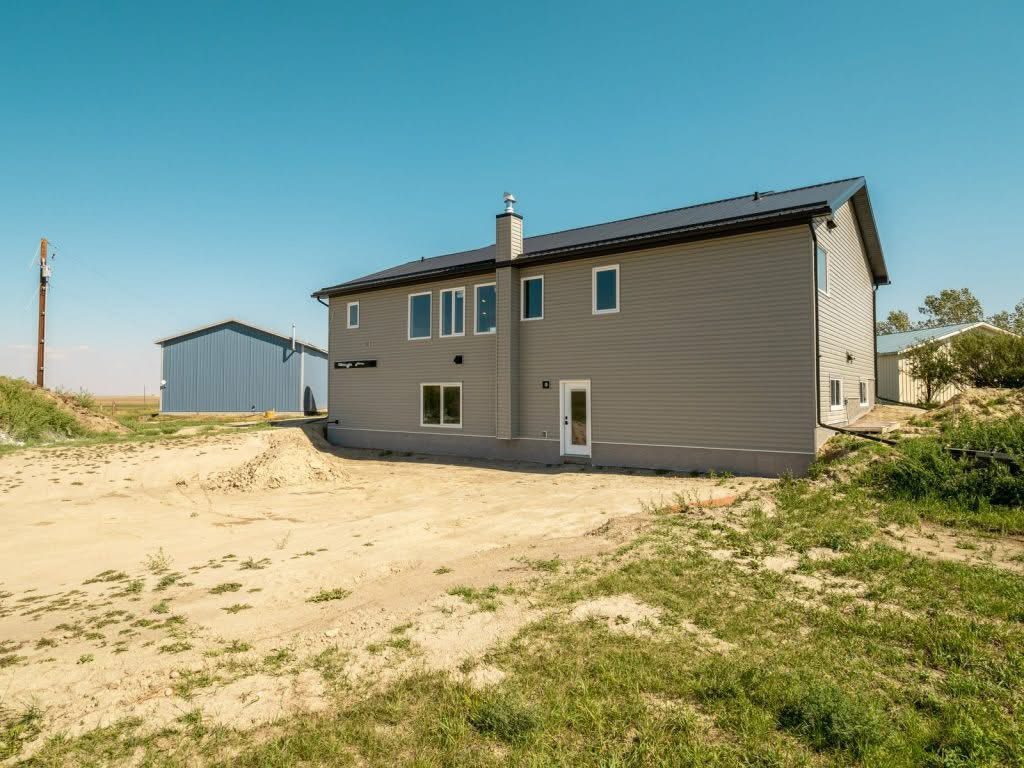 A two-story, beige house sits on a sandy terrain under a clear blue sky. Nearby, a large blue barn adds to the open rural ambiance.