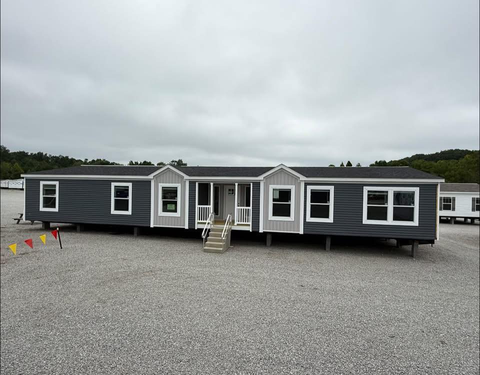 A large, gray modular home with white trim sits on gravel under an overcast sky. Red and yellow pennant flags mark the area, conveying a display setting.