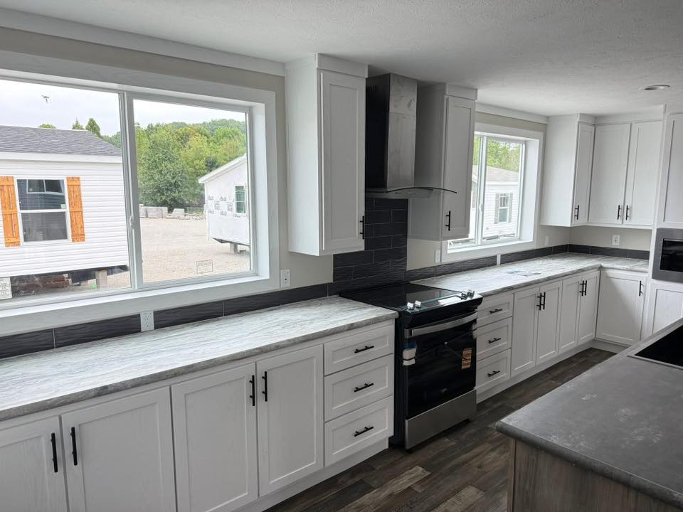 Modern kitchen with white cabinets, gray countertops, and stainless steel appliances. Large windows provide natural light, creating an airy, fresh atmosphere.