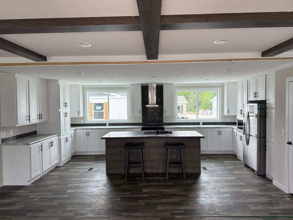 Modern kitchen with white cabinets, dark countertops, and an island with two stools. Large windows provide natural light, creating an airy feel.