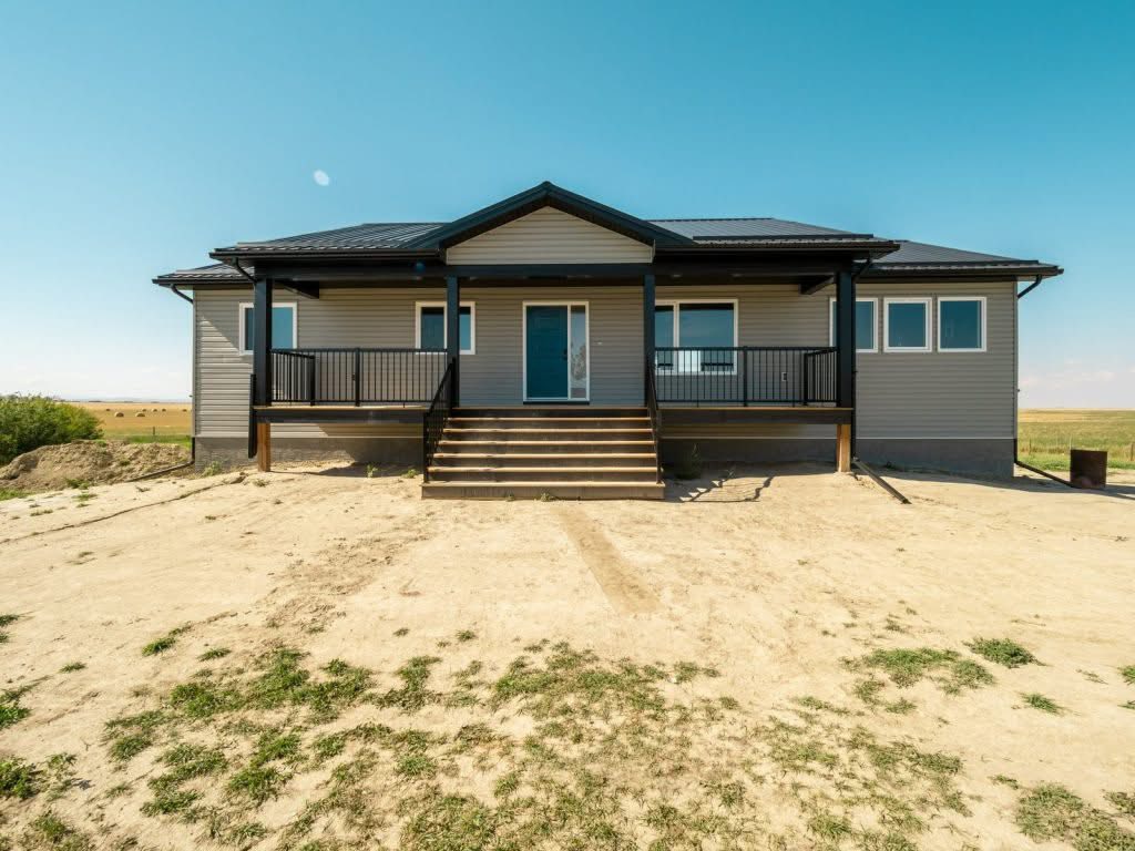 Single-story house with gray siding and black roof sits in a barren landscape under a clear blue sky. Wooden steps lead to a central porch.
