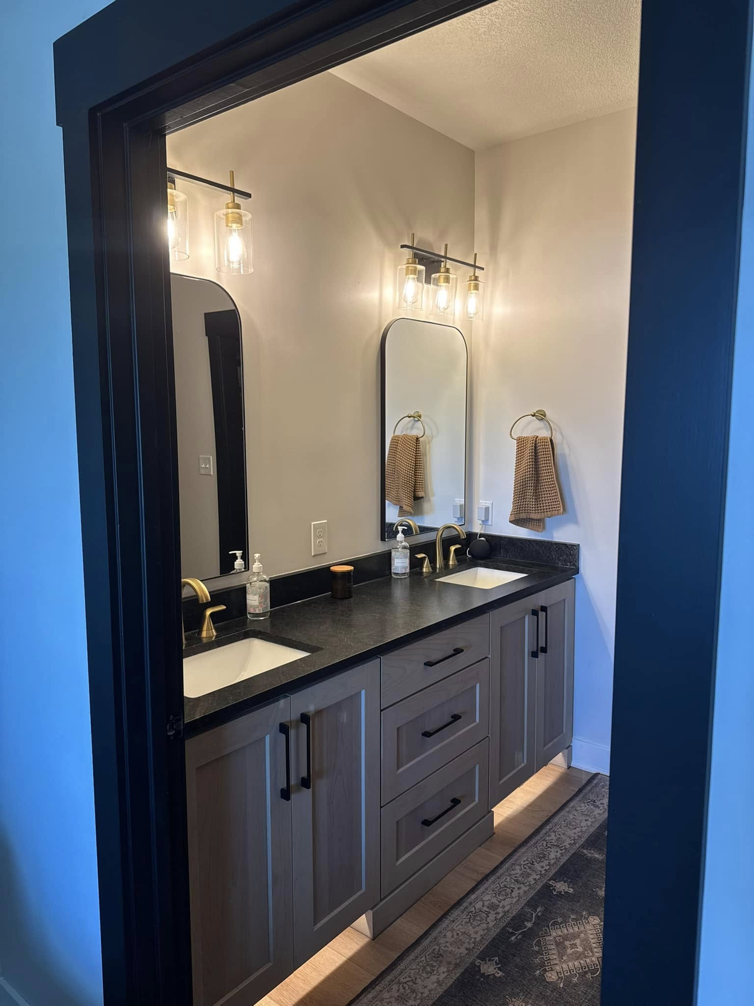 Modern bathroom with a double vanity, wood cabinets, black countertop, and gold fixtures. Two mirrors and warm lights create a cozy, elegant feel.