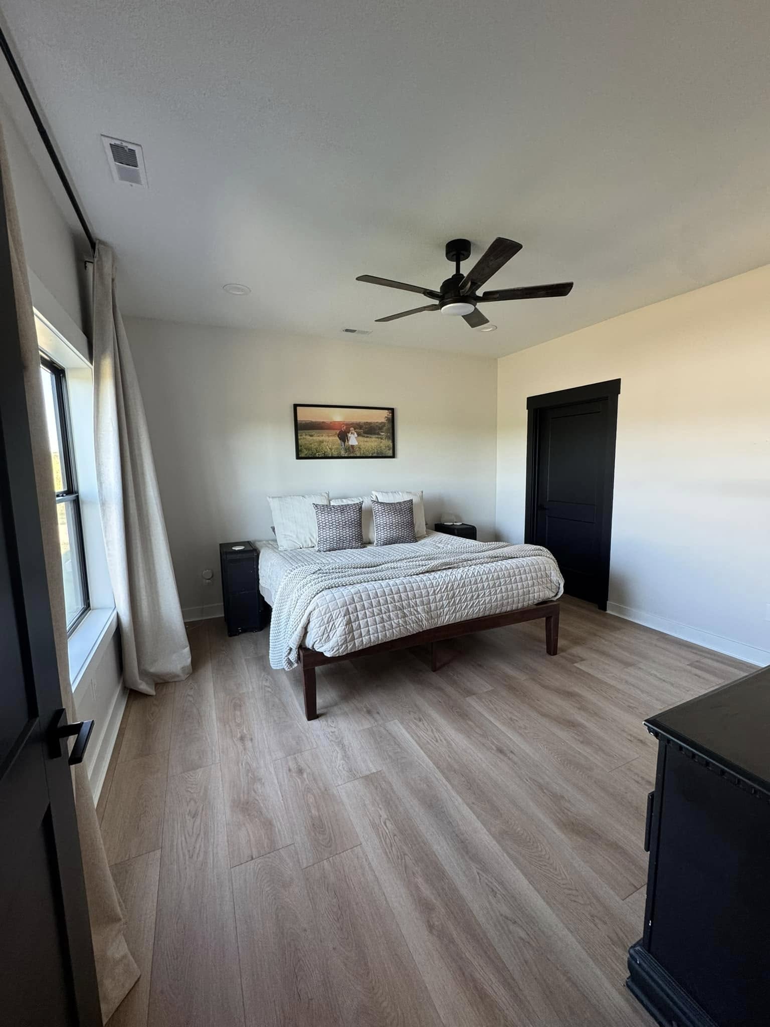 A minimalist bedroom with light wood flooring features a neatly made bed with a beige quilt, two nightstands, a ceiling fan, and a landscape painting.