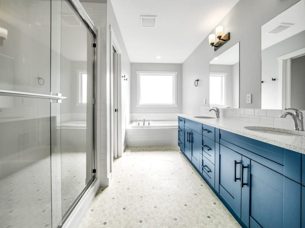 Bright, modern bathroom with a blue vanity, dual sinks, and large mirrors. Glass shower on the left, bathtub by a window, and elegant lighting.