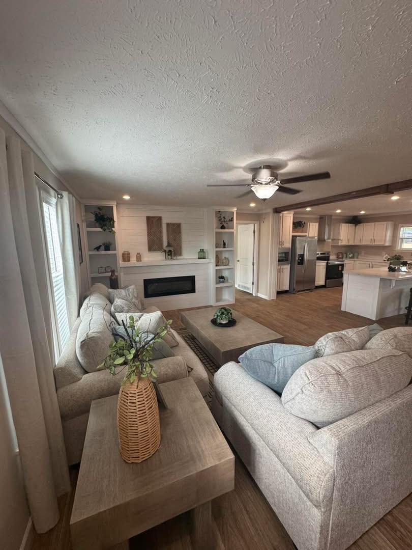 Stylish living room with beige sofas, wooden coffee table, and ceiling fan. Fireplace and decorative shelves on back wall. Kitchen visible in background. Cozy atmosphere.