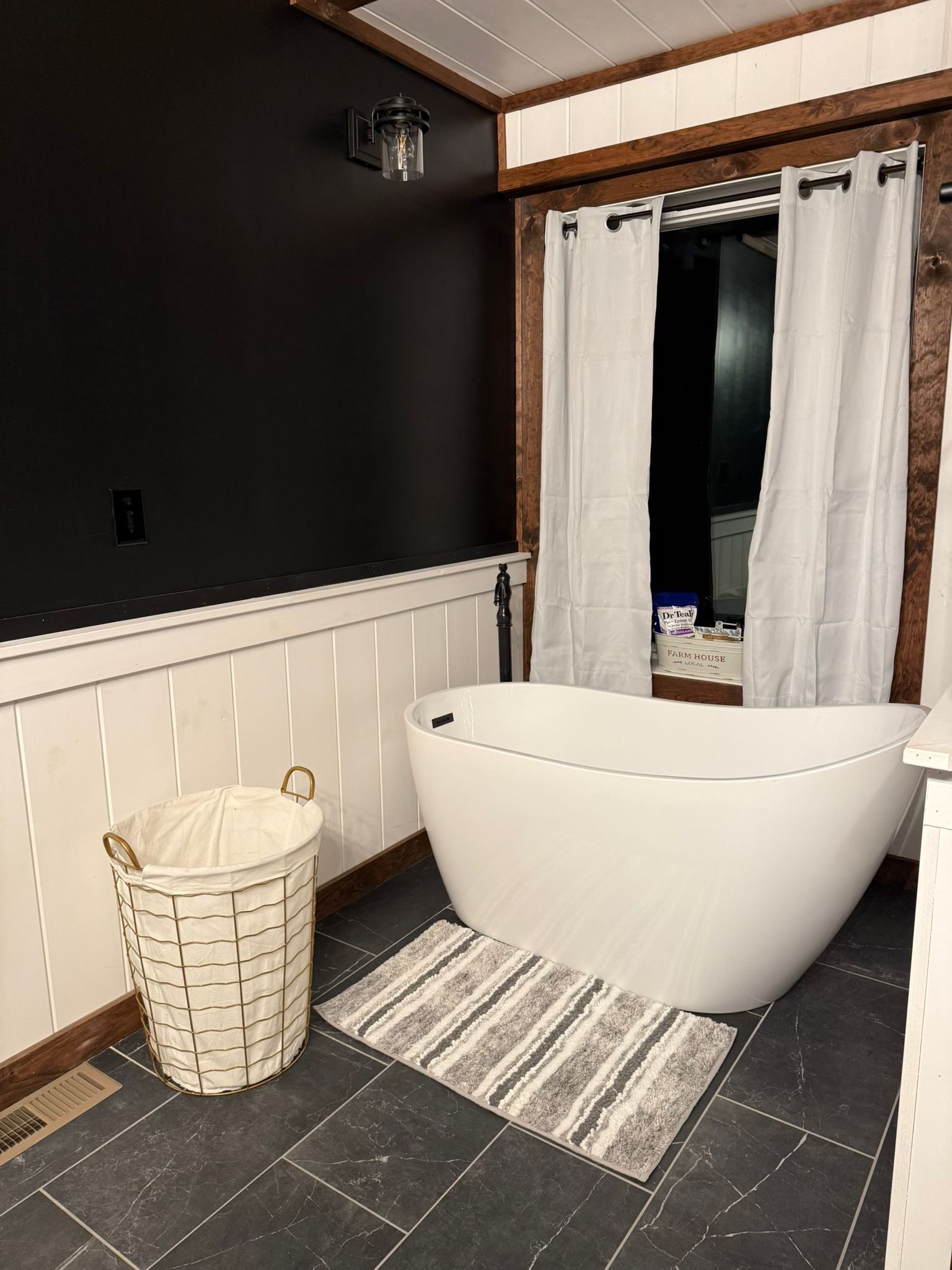 Modern bathroom with a deep white freestanding tub on gray tiles. A striped rug lies nearby, with a wire laundry basket accented in gold. White curtains cover a large window, set within dark brown wood trim. The upper black wall contrasts with the lower white paneling, creating a minimalist yet cozy atmosphere.