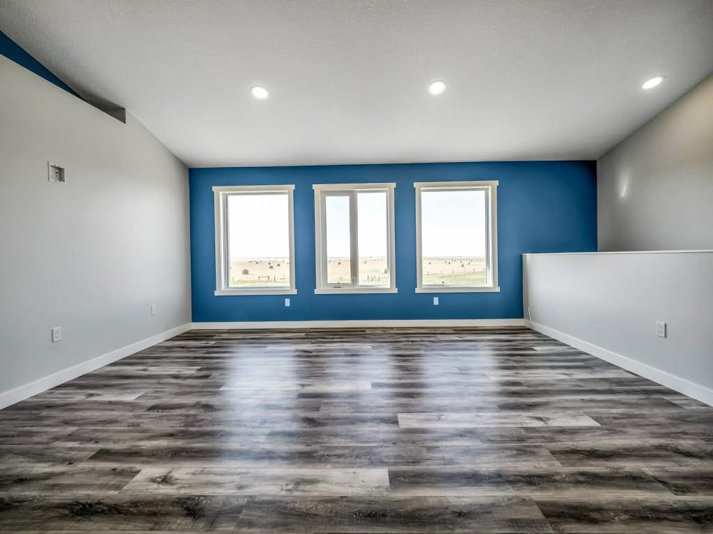 Spacious room with a blue accent wall and three large windows offering a view of an open landscape. Modern gray wood flooring, white walls, serene ambiance.