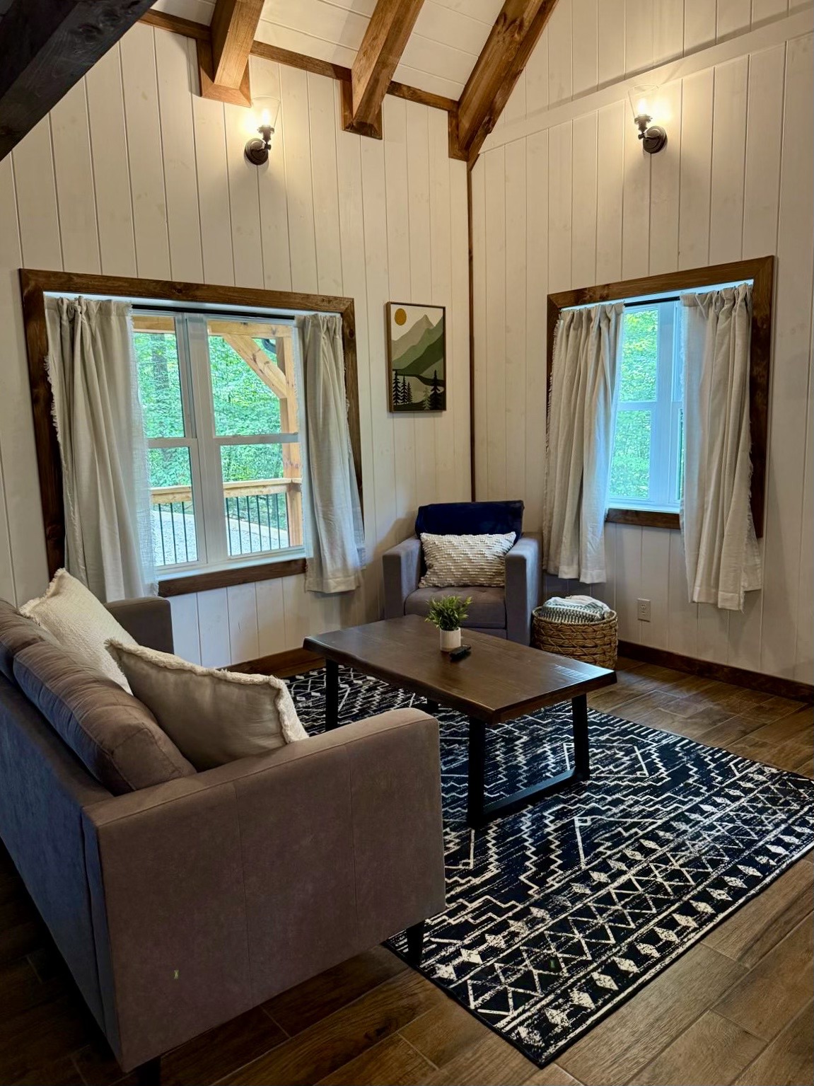 Cozy living room with a wooden floor, white paneled walls, and exposed beams. Features a gray sofa, armchair, and patterned rug, creating a rustic yet inviting atmosphere.