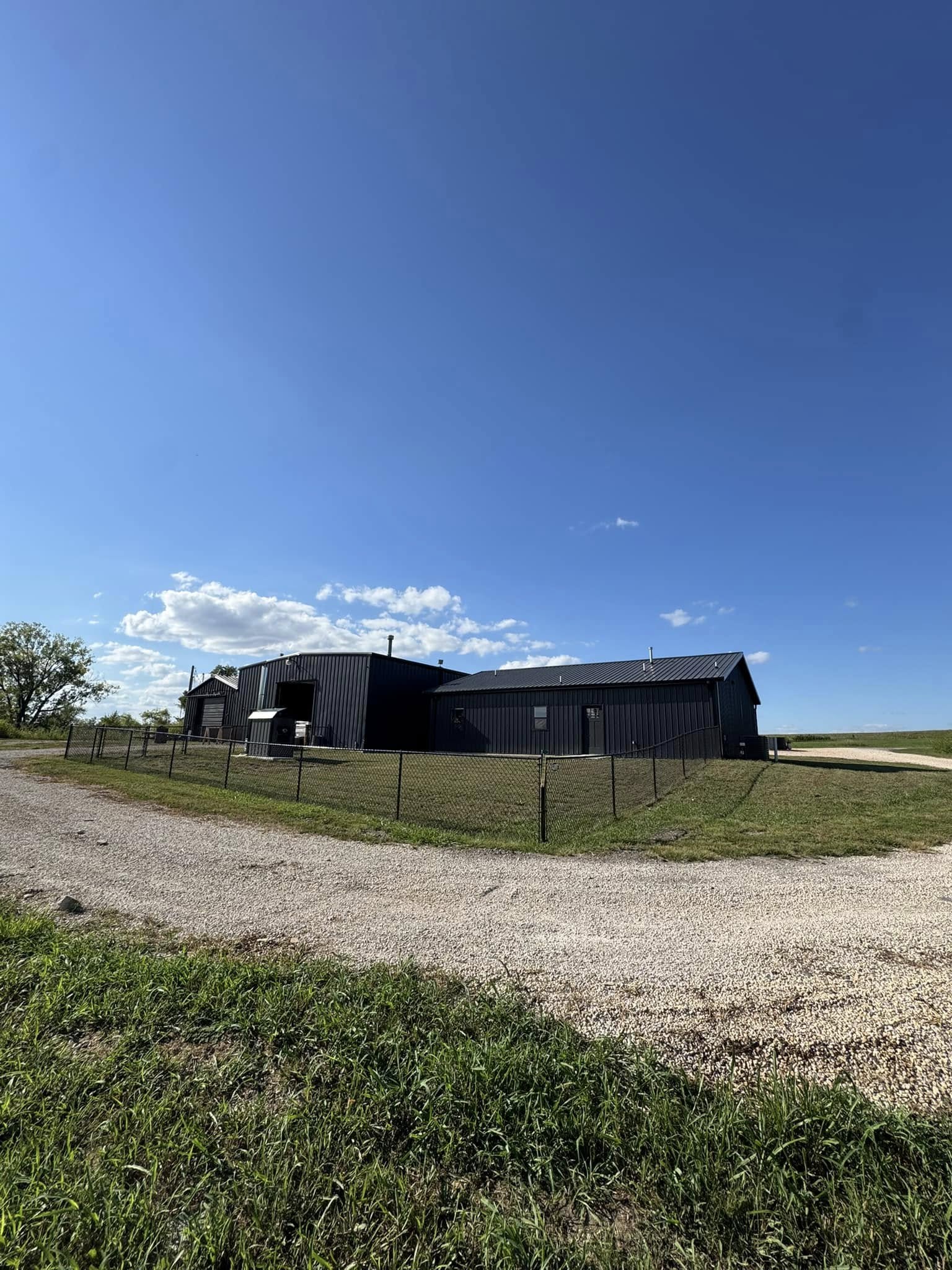 Large black farmhouse under clear blue sky with scattered clouds. Surrounded by a dirt path and green grass, creating a peaceful rural setting.